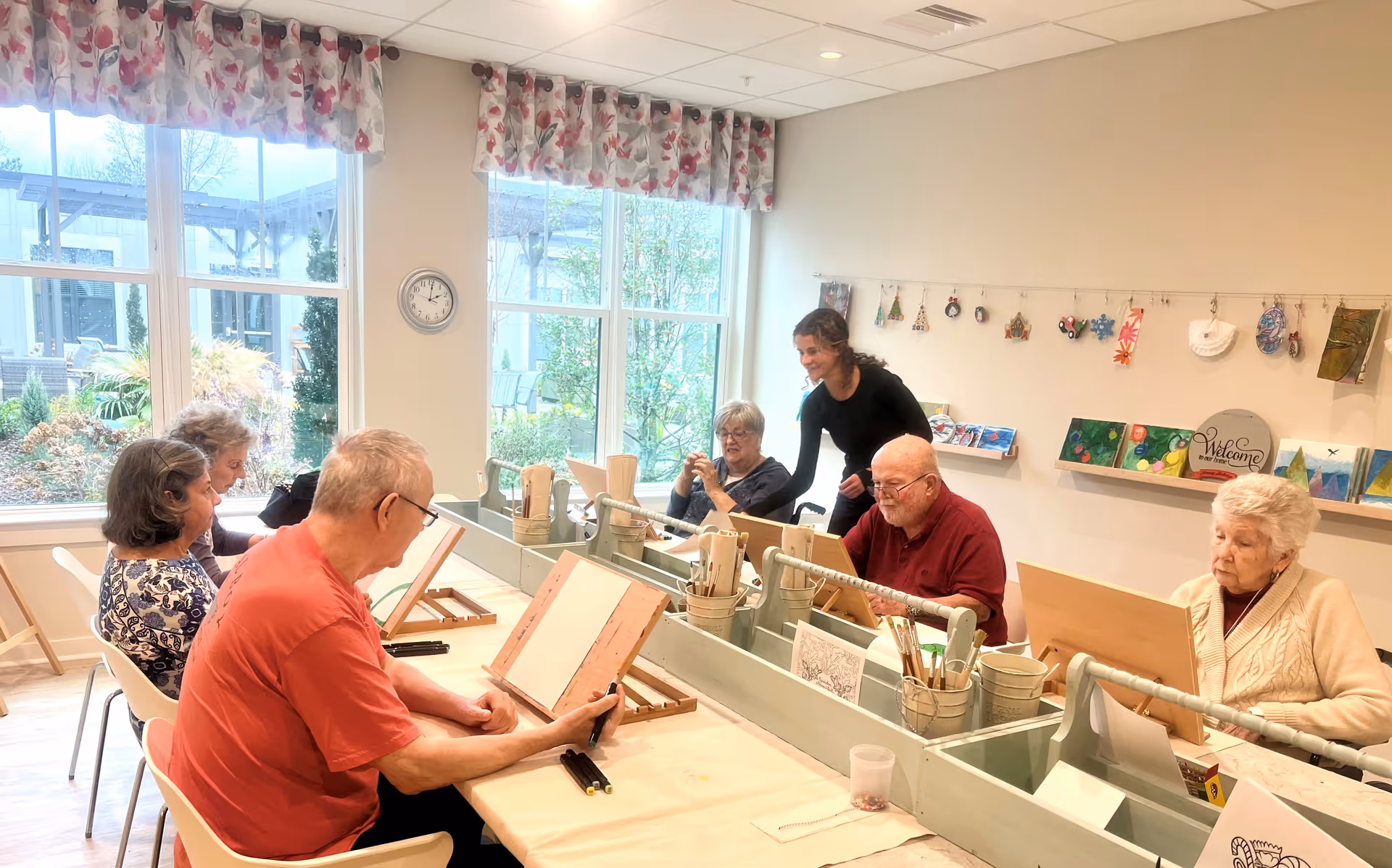 A group of elderly people seated around a table in a bright room with large windows, engaging in an art activity with drawing boards and art supplies. A caregiver or instructor is standing and assisting one of the participants. The room has floral curtains, a clock on the wall, and various artworks displayed on shelves and hanging on a string.
