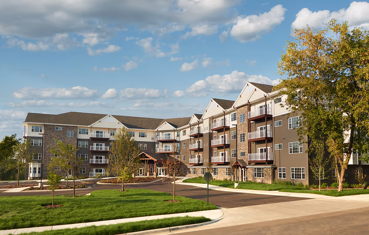 Exterior view of a modern four-story senior living facility building with balconies, surrounded by green lawns, trees, and a clear blue sky with scattered clouds.