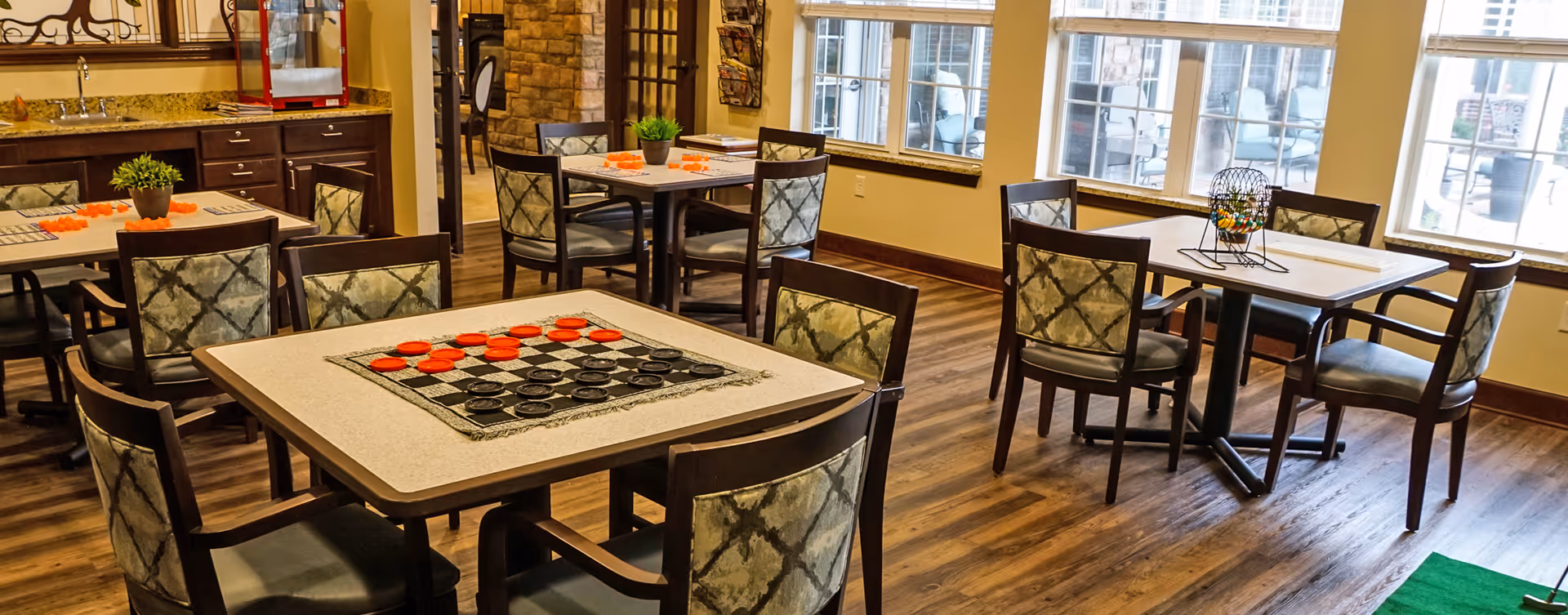 A well-lit common area with several tables and chairs arranged on a wooden floor. One table has a checkerboard game set up with red and black pieces, while another table has a bingo cage with numbered balls. Large windows let in natural light, and there is a countertop with a sink and cabinets in the background.