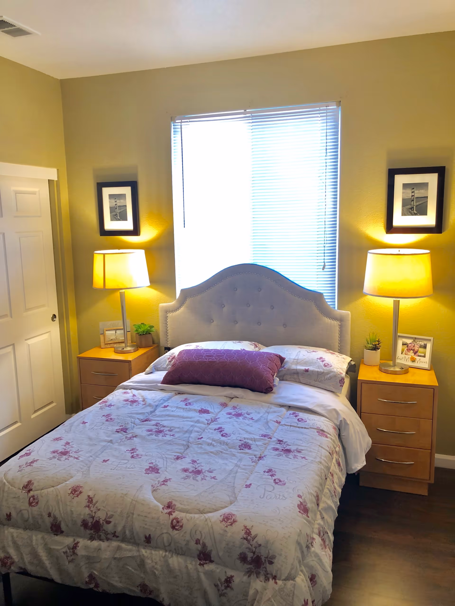 A cozy bedroom with a bed centered against a wall with a window covered by blinds. The bed has a light-colored upholstered headboard and is made with a floral-patterned comforter and a purple decorative pillow. On each side of the bed are wooden nightstands with three drawers, each topped with a lit table lamp, a small plant, and a framed photo. Two black-and-white framed pictures hang on the wall above the nightstands.