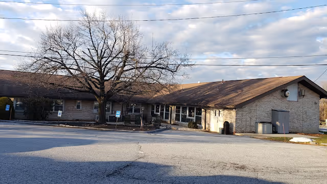 Exterior view of a single-story brick building with a brown roof, a large leafless tree in front, and a paved parking area with handicap parking signs.