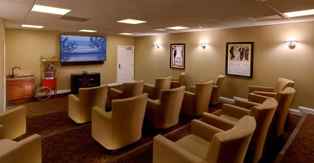 Small senior-living screening room with rows of beige armchairs facing a wall-mounted TV, a popcorn machine, and framed posters.