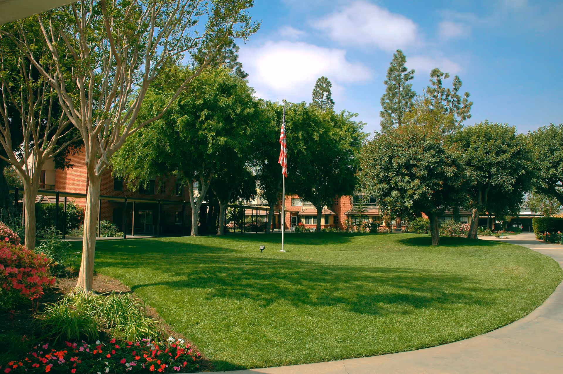A well-maintained outdoor garden area with green grass, trees, and colorful flowers. There is an American flag on a flagpole in the center of the lawn, and a brick building is visible in the background under a partly cloudy blue sky.