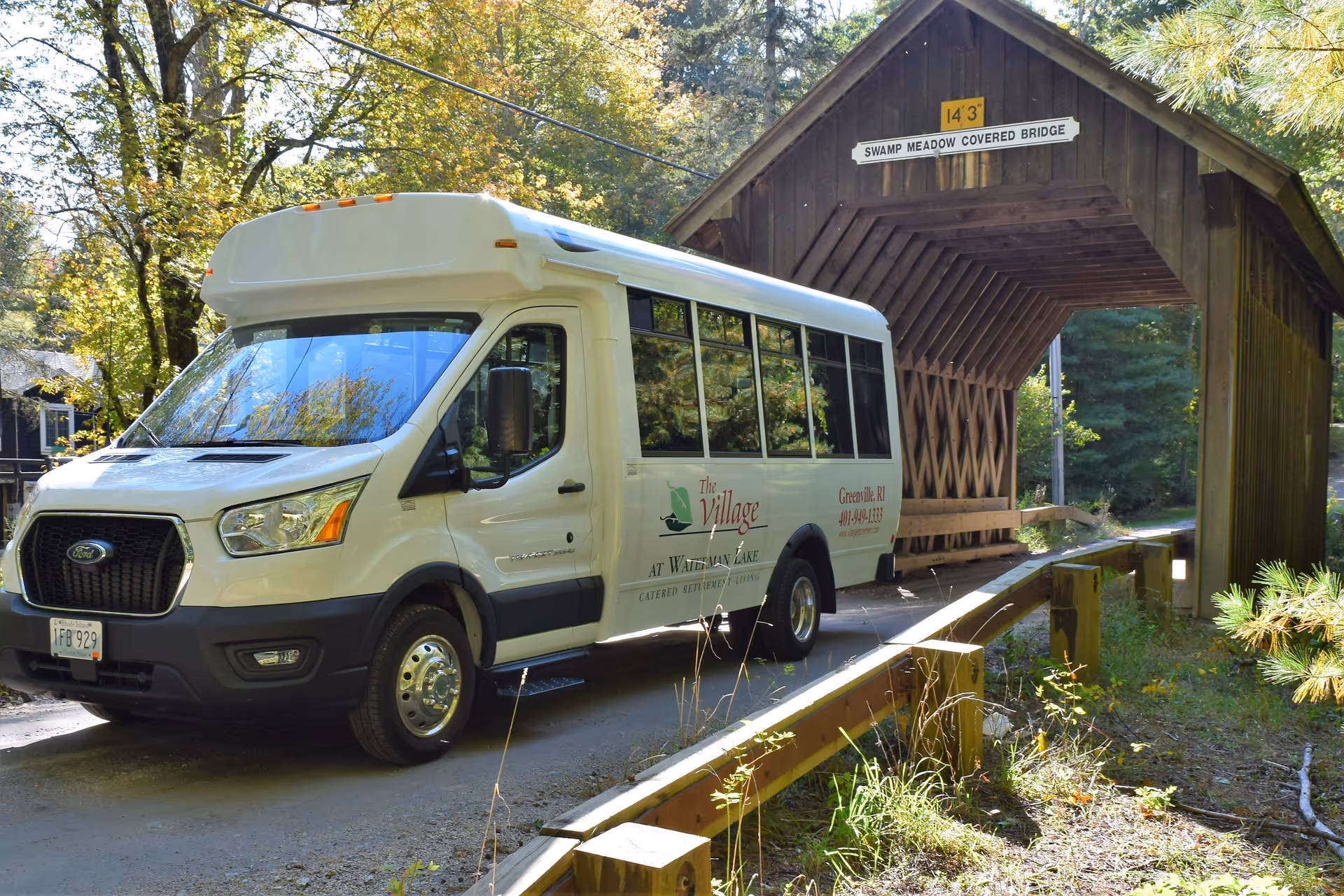 A white shuttle bus from The Village at Waterman Lake is parked near the entrance of a wooden covered bridge labeled 'Swamp Meadow Covered Bridge' surrounded by trees and greenery on a sunny day.
