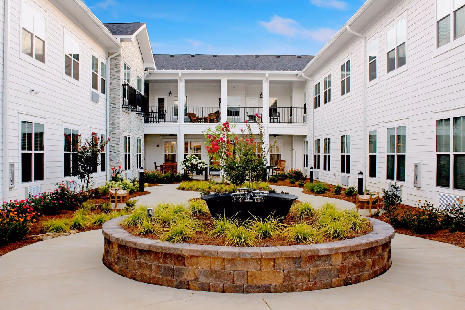 A beautifully landscaped courtyard at The Blake at Carnes Crossroads featuring a circular stone planter with a fountain, surrounded by colorful flowers and greenery, with buildings in the background.