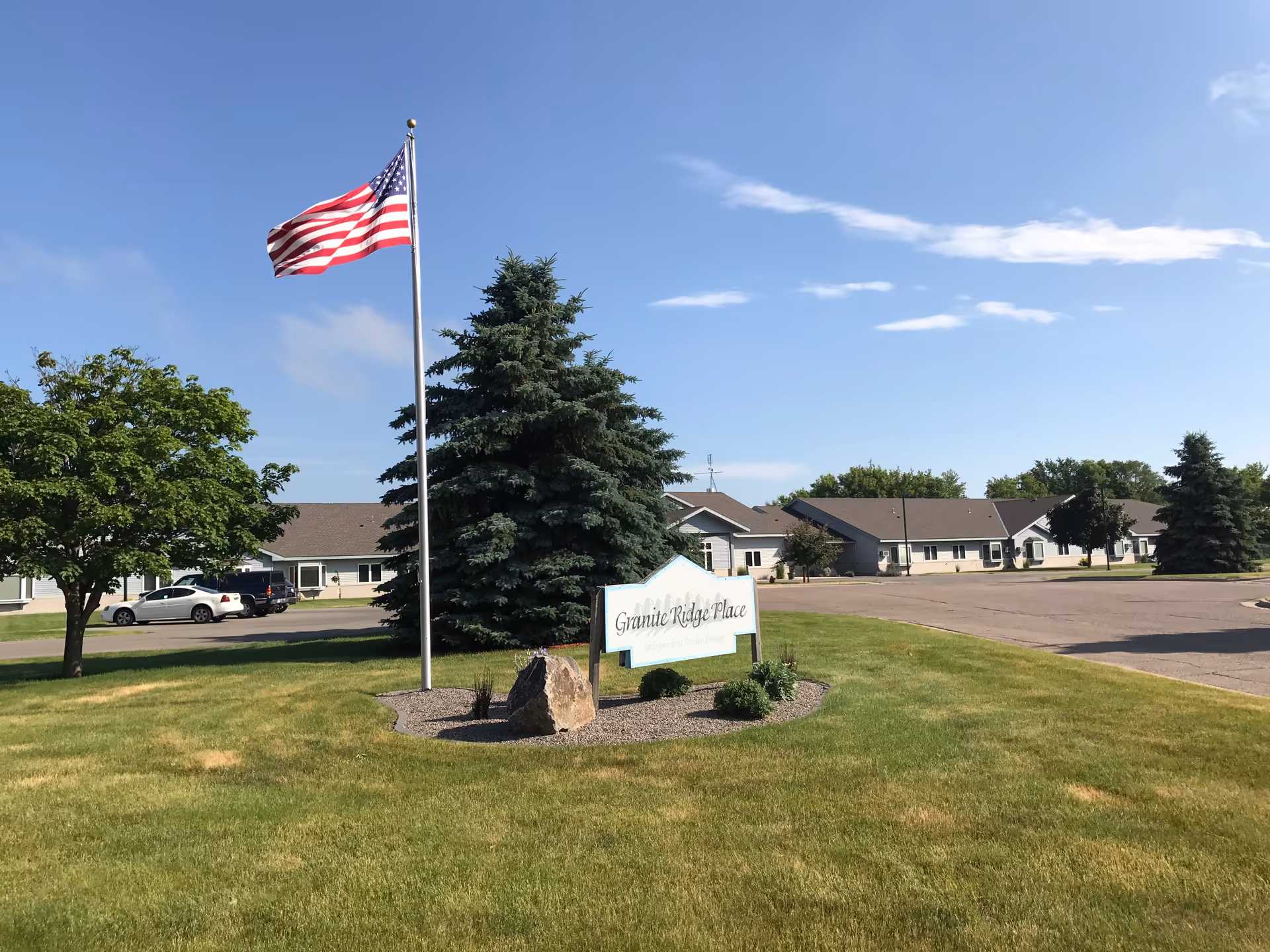 Exterior view of Granite Ridge Place senior living facility with a flagpole, sign, trees, and a parking lot.