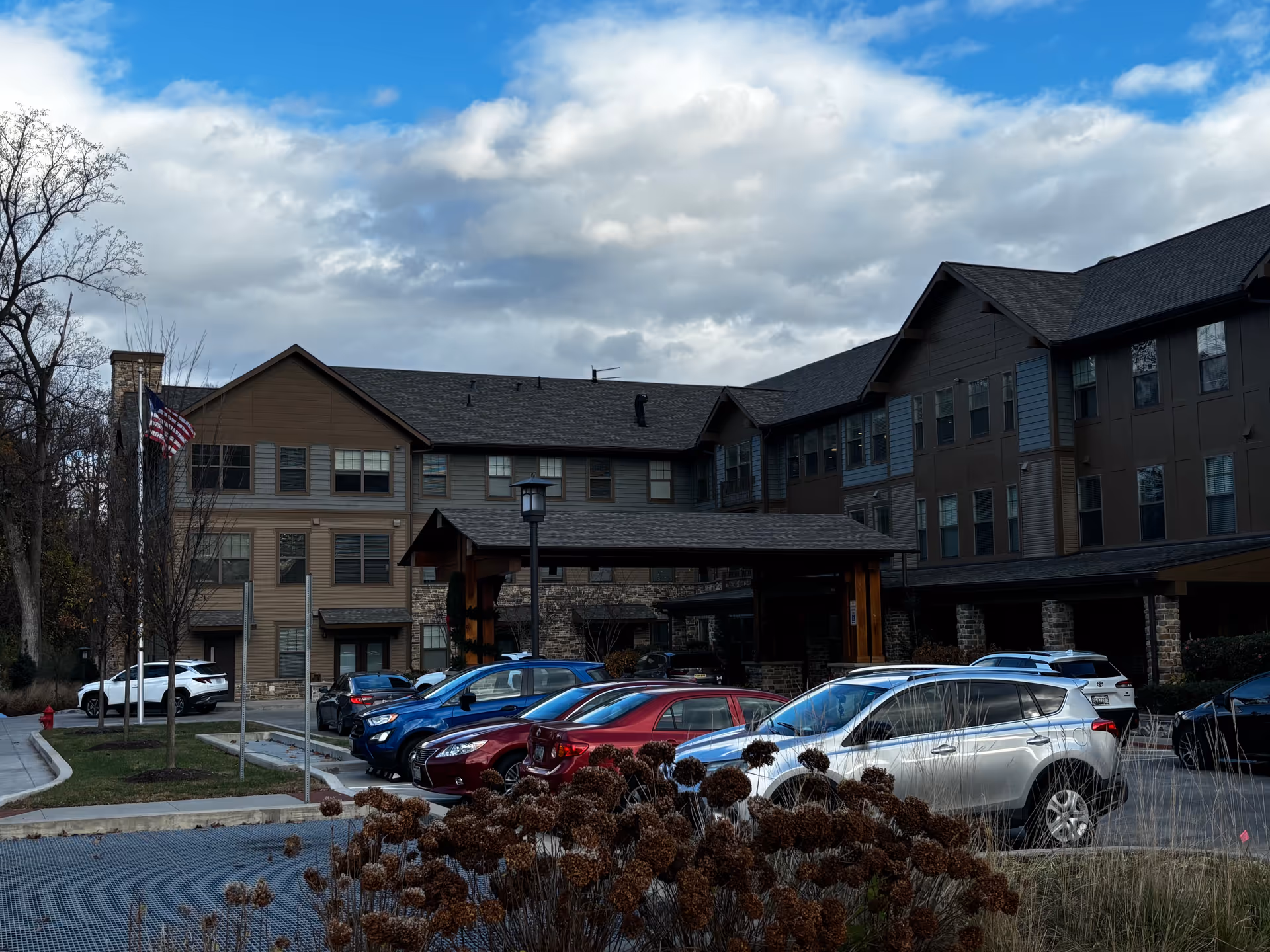 Exterior view of a senior living facility building with multiple floors, a covered entrance, several parked cars in front, an American flag on a flagpole, and some dried plants in the foreground under a partly cloudy sky.