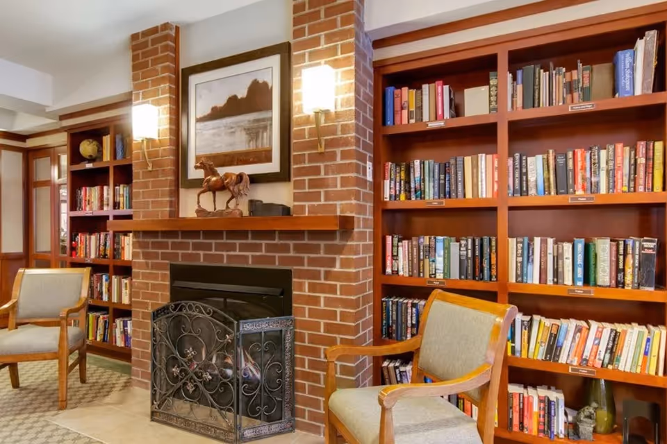 Cozy interior room with a brick fireplace in the center, flanked by wooden bookshelves filled with books. Two wooden armchairs with light green cushions are positioned near the fireplace. Above the fireplace is a framed picture and a horse sculpture on the mantel. The room is warmly lit by wall sconces.