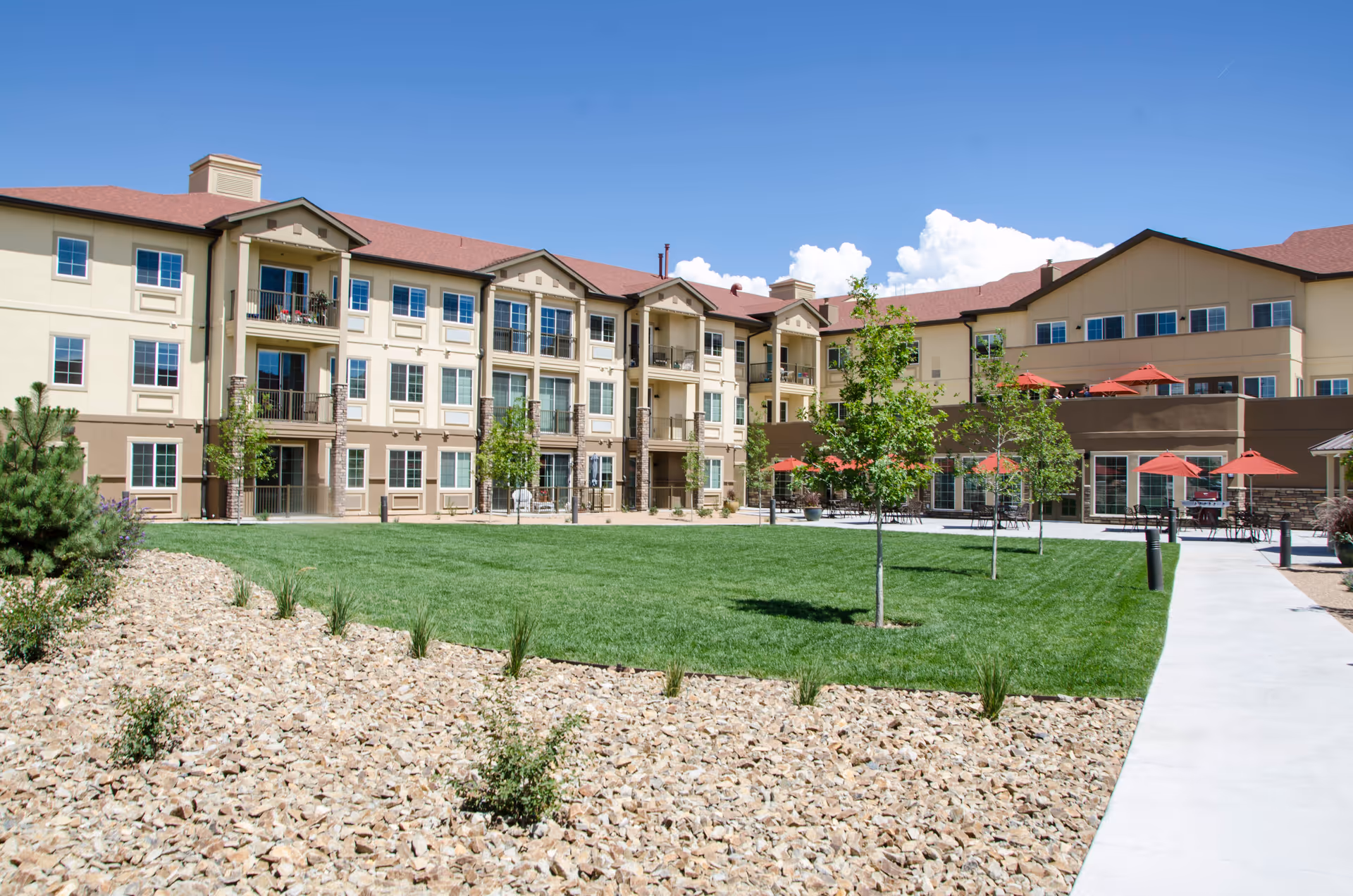Exterior view of Palmilla Senior Living facility showing a three-story building with balconies and numerous windows. In front of the building is a well-maintained green lawn with young trees, surrounded by a rock garden and a paved walkway. Several red patio umbrellas and outdoor seating are visible on the right side under a clear blue sky.