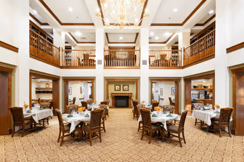 A spacious dining room with multiple tables covered in white tablecloths and set with green napkins and floral centerpieces. The room features a high ceiling with wooden beams, a chandelier, and a fireplace at the far end. There is a second-floor balcony with wooden railings overlooking the dining area.