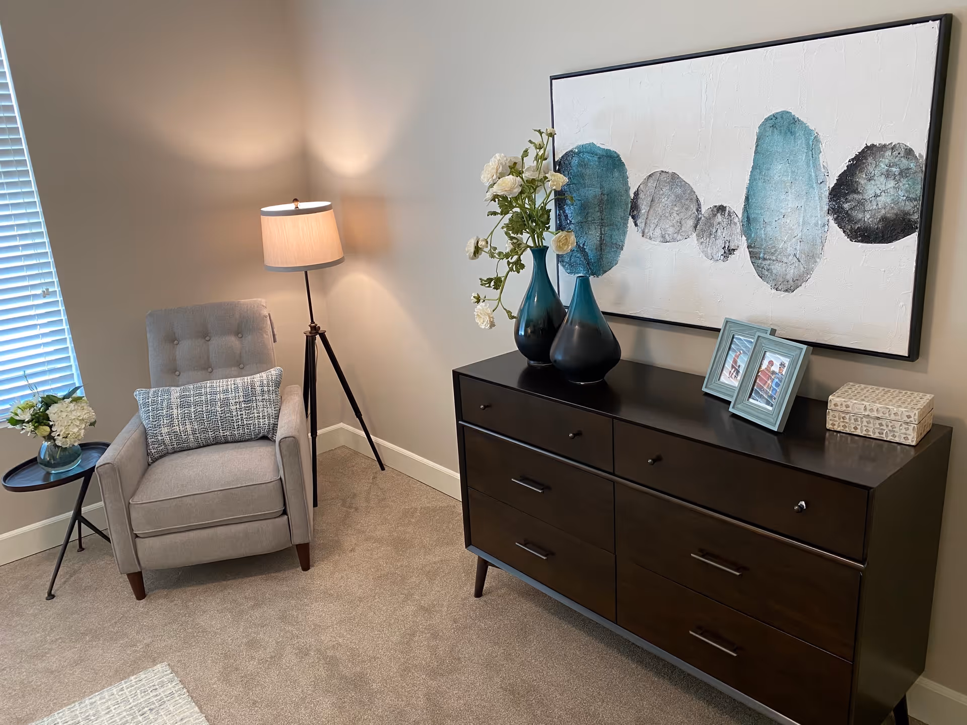A cozy corner of a room featuring a gray upholstered armchair with a blue patterned lumbar pillow, a small round side table with a floral arrangement, a tripod floor lamp with a beige shade, and a dark wooden dresser topped with two blue-green vases holding white flowers, two framed photos, and a decorative box. Above the dresser hangs a large abstract painting with blue and gray circular shapes on a white background.