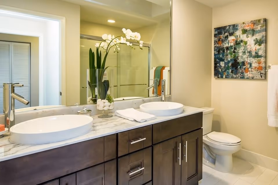 Bright modern bathroom with dual vessel sinks on a marble vanity, large mirror, glass shower, and a toilet.