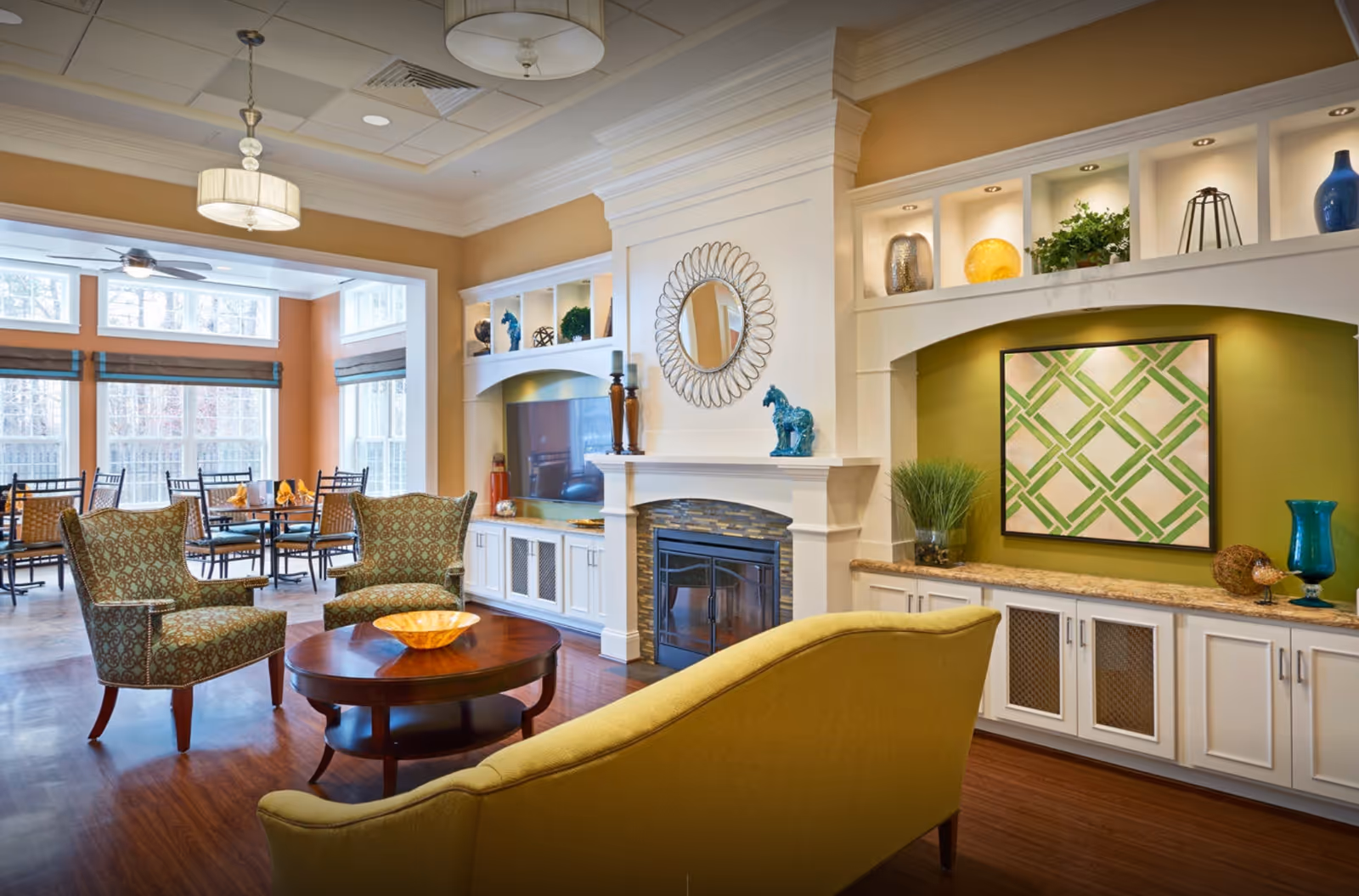 A cozy and well-decorated living room area in a senior living facility featuring a yellow sofa, two patterned armchairs, a round wooden coffee table with a decorative bowl, a fireplace with a circular mirror above it, built-in shelves with decorative items, and a dining area with tables and chairs near large windows letting in natural light.