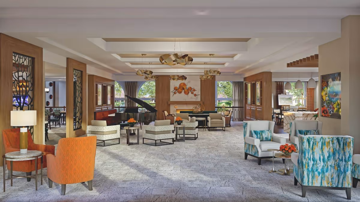 A spacious and well-lit senior living facility common area with modern and comfortable seating arrangements including orange, patterned, and blue-green upholstered chairs around small tables. The room features decorative ceiling lights, a grand piano, large windows with views of greenery outside, and artwork on the walls. There is a fireplace in the center back wall with a floral painting above it.
