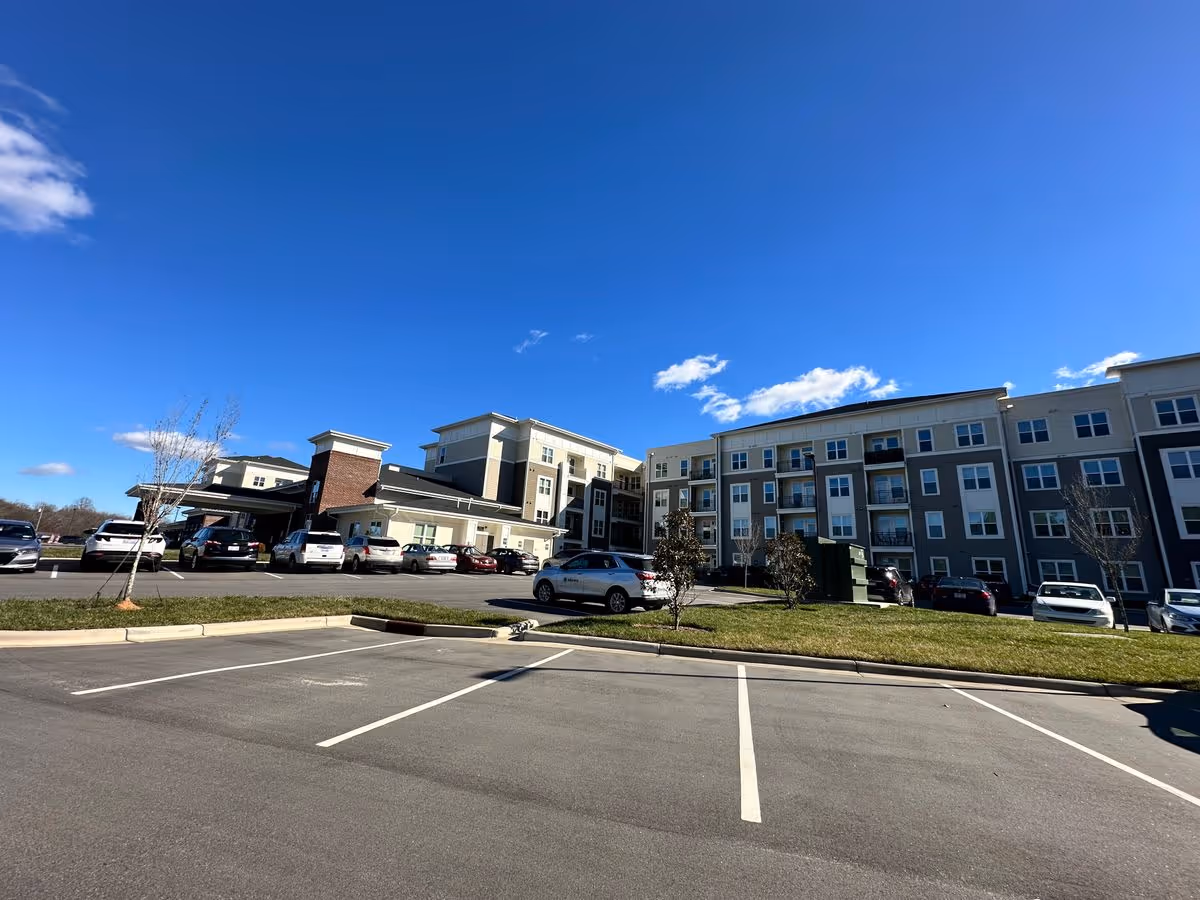 Front exterior of a multi-story senior living building with a parking lot and cars under a clear blue sky.