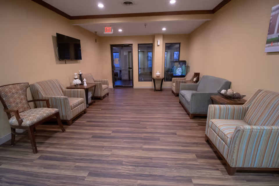 A waiting or sitting area with multiple upholstered chairs and sofas arranged along the walls. There is a flat-screen TV mounted on the left wall, small side tables with decorative items, and an aquarium on the right side. The room has wood flooring, beige walls, and a ceiling with recessed lighting and crown molding. A glass door and windows are visible at the far end of the room.