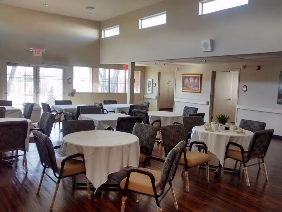 A dining room with several round and rectangular tables covered with white tablecloths, surrounded by cushioned chairs. The room has large windows letting in natural light, wooden flooring, and light-colored walls with framed artwork. There are exit signs above the doors and some table settings including cups and a small plant on one table.