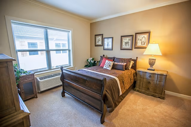 A cozy bedroom with a wooden bed frame, a brown and beige bedspread, and multiple pillows. There is a wooden nightstand with a lamp on the right side of the bed, and three framed pictures hanging on the beige wall above the bed. A large window with blinds lets in natural light, and there is an air conditioning unit below the window. The room has beige carpet and additional wooden furniture pieces.