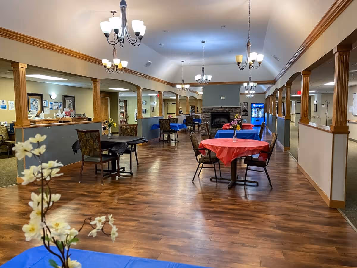A spacious dining area in an assisted living facility with multiple round tables covered with red, blue, and black tablecloths. Each table has a small vase with flowers. The room features wooden flooring, wooden columns, and several chandeliers hanging from a vaulted ceiling. A stone fireplace is visible at the far end, and a Pepsi vending machine is near the back wall.