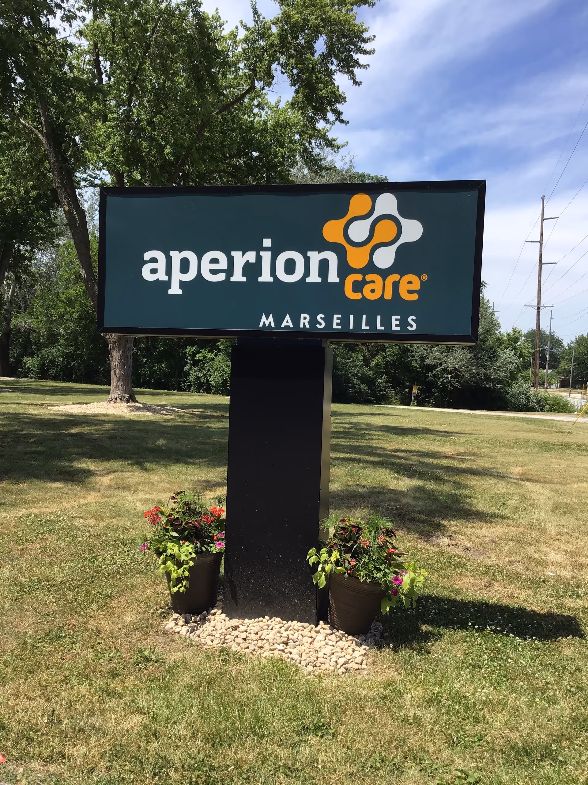 A freestanding sign reading "Aperion Care Marseilles" on a grassy lawn with potted flowers and trees in the background.