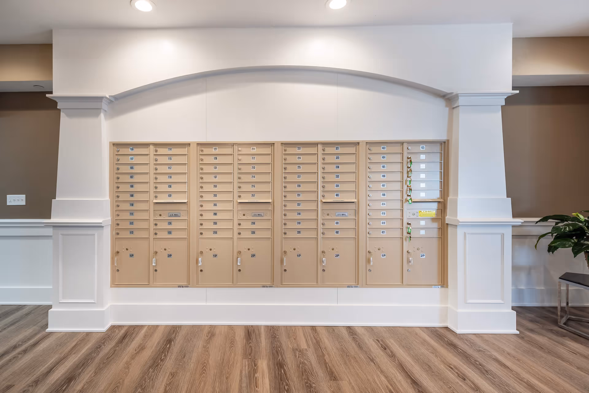 A wall of beige mailboxes with numbered compartments in a senior living facility. The mailboxes are set within a white wall with decorative molding and an arch above. The floor is wood with a natural finish, and there is a small plant and a bench visible on the right side.