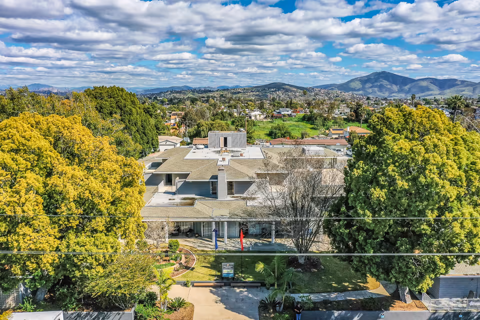 Aerial view of Monte Vista Village facility surrounded by large green trees with a mountainous landscape in the background under a partly cloudy sky.