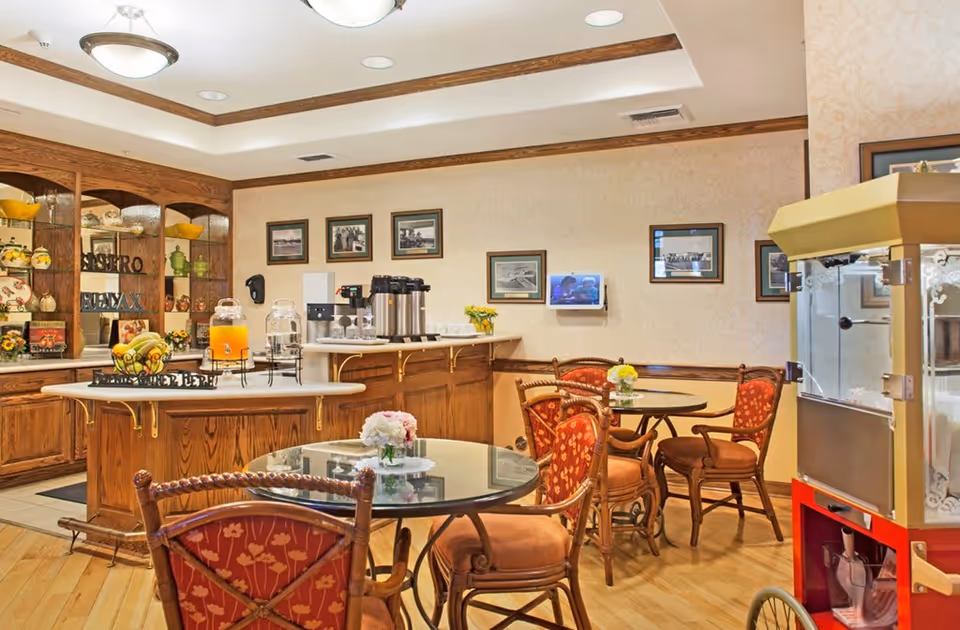 A cozy dining area in a senior living facility with round glass-top tables and cushioned wooden chairs. The room features a wooden island with beverage dispensers, coffee urns, and a bowl of fruit. The walls are decorated with framed pictures, and there is a popcorn machine on the right side. The space has warm lighting and wooden flooring.