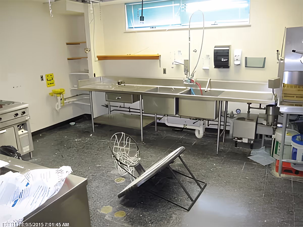 An industrial kitchen area with stainless steel counters and sinks. There is a large metal table or rack tipped over on the floor. The room has a window with blue blinds, shelves on the wall, and various kitchen equipment including a stove and a dishwasher. The floor appears to be dark and textured.