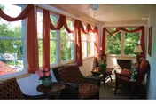 Sunlit enclosed sitting room with upholstered chairs, small tables, potted plants, and large windows draped with red valances.