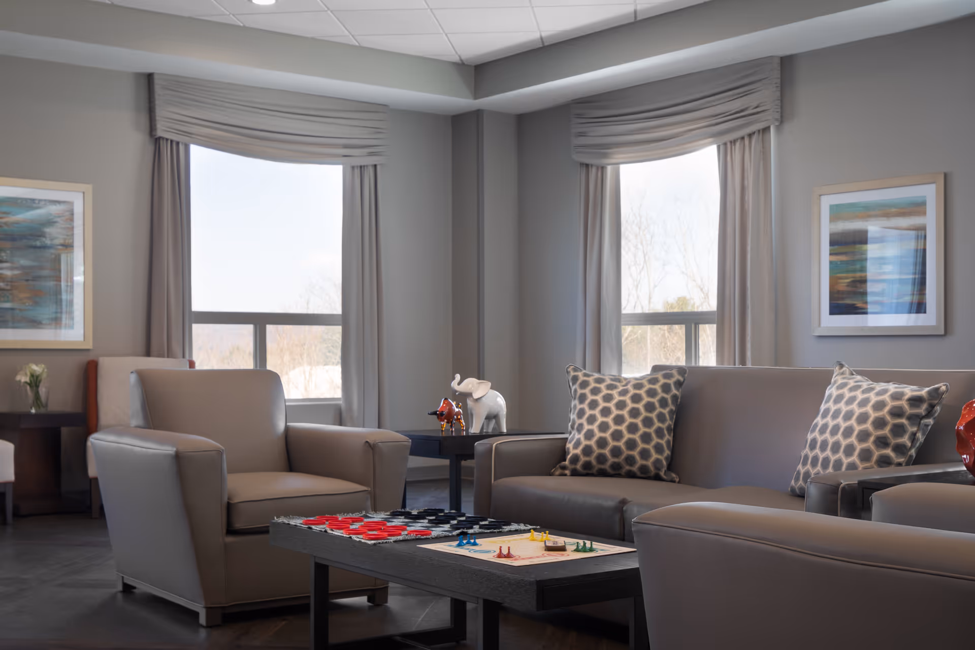 A cozy living room area with gray leather armchairs and a sofa adorned with patterned pillows. A coffee table in the center holds a board game with colorful pieces. Two large windows with light gray curtains allow natural light to fill the room. The walls are decorated with framed abstract artwork, and small decorative items, including a white elephant figurine, are placed on side tables.