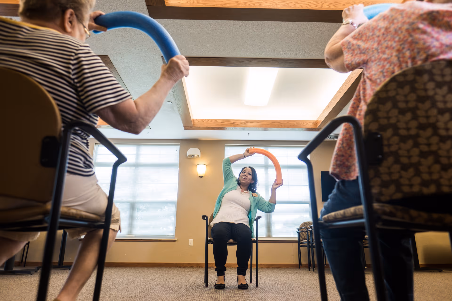 A group of elderly individuals seated in chairs participating in a seated exercise class led by an instructor holding a blue exercise noodle in a well-lit room with large windows.