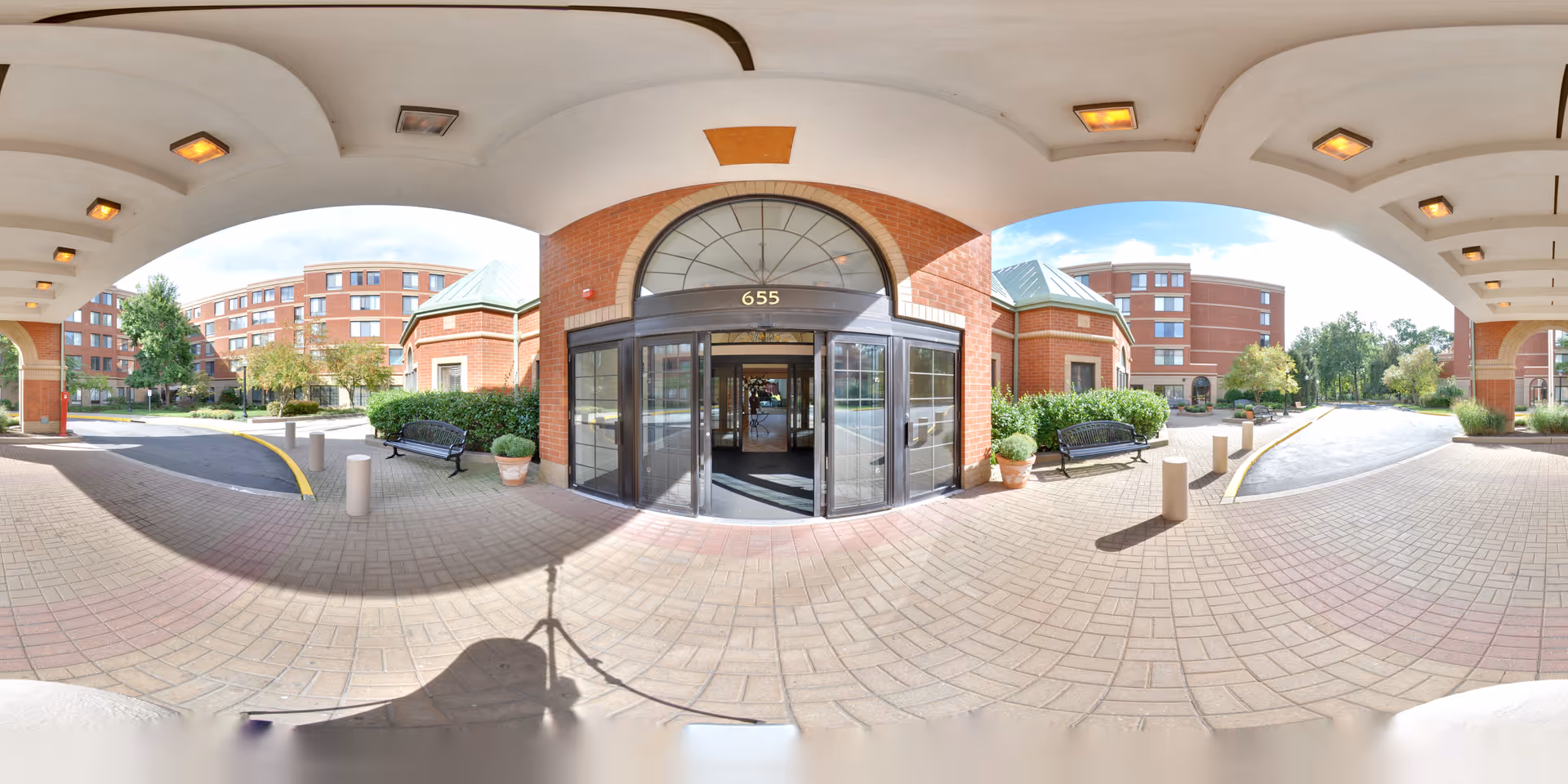 Entrance area of Five Star Premier Residences of Teaneck showing a covered driveway with brick pillars and a glass door entrance marked with the number 655. There are benches, potted plants, and a multi-story brick building in the background under a partly cloudy sky.