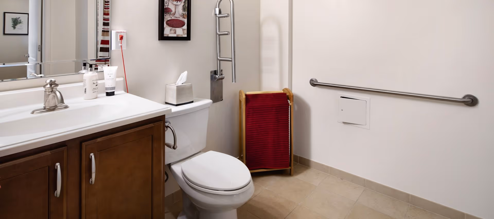Accessible bathroom with a toilet, vanity sink and mirror, red towel rack, and a wall-mounted grab bar.