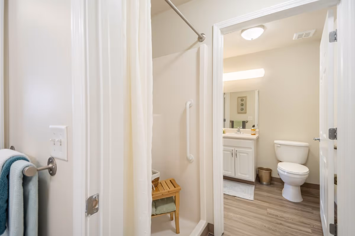 View of a clean bathroom featuring a white toilet, a vanity with a sink and mirror, a shower area with a white curtain and a wooden bench inside, and a towel rack with folded towels on the left wall.