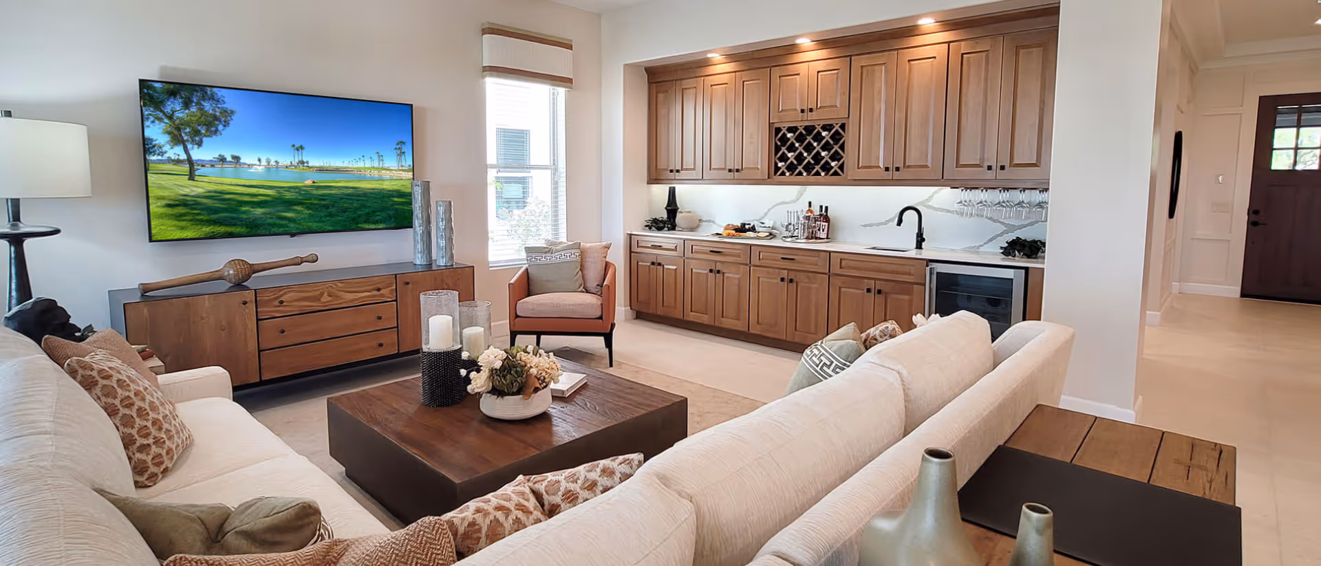 Bright open living area featuring a cream sectional sofa, wooden coffee table, wall-mounted TV, and a long wooden wet bar with cabinetry.