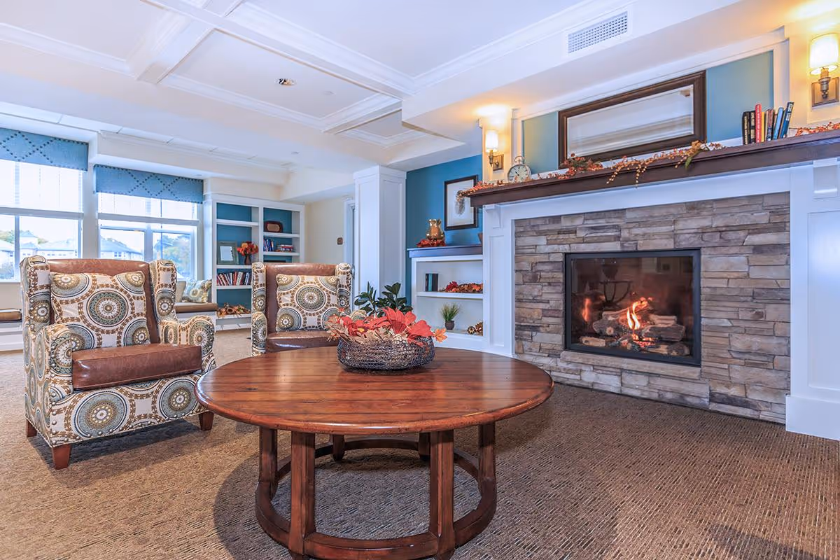 A cozy living room area in an assisted living facility featuring two patterned armchairs with brown leather cushions, a round wooden coffee table with a decorative centerpiece, a stone fireplace with a fire burning, built-in white shelves with books and decorations, and large windows with blue valances letting in natural light.