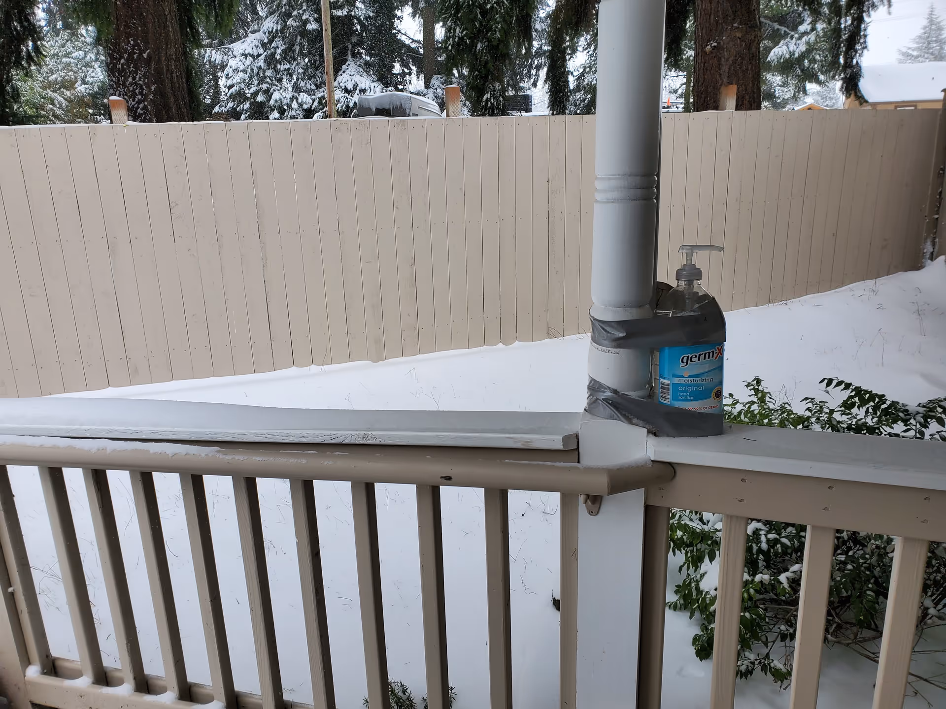 A snowy outdoor scene showing a beige wooden fence and a railing with a white post. A bottle of Germ-X hand sanitizer is taped to the post with gray duct tape. Snow covers the ground and some green bushes are visible near the fence.