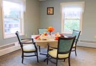 Small dining room with a round table set for four, surrounded by chairs and two windows with sheer curtains.