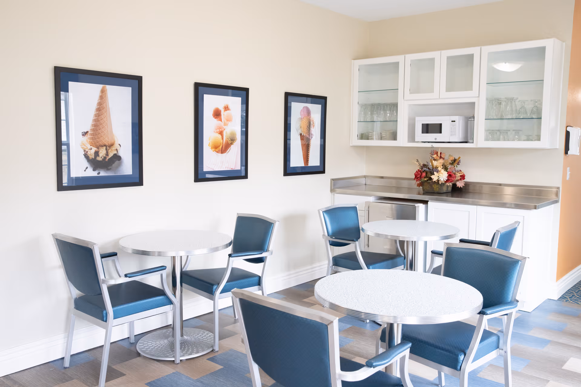 A small dining area with three round tables and blue cushioned chairs. On the wall are three framed pictures of ice cream cones and scoops. There is a white cabinet with glass doors containing glassware and a microwave on the countertop, along with a floral arrangement.
