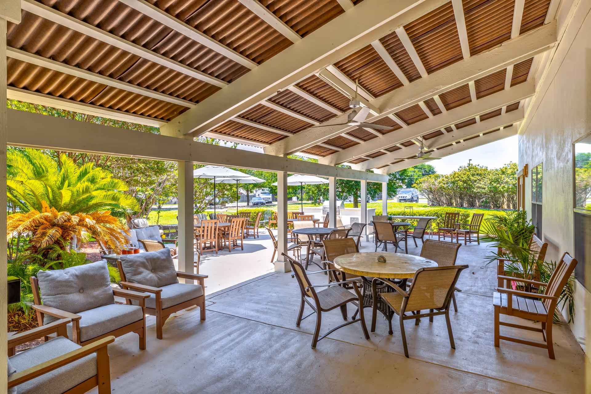 Covered outdoor patio area with multiple seating arrangements including cushioned armchairs, round tables with chairs, and wooden benches. The patio overlooks a garden with green plants and trees, and there are umbrellas providing shade over some tables in the background.