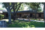 Single-story brick building front with mature trees, a covered entryway, walkways, lawn, and a small fountain.