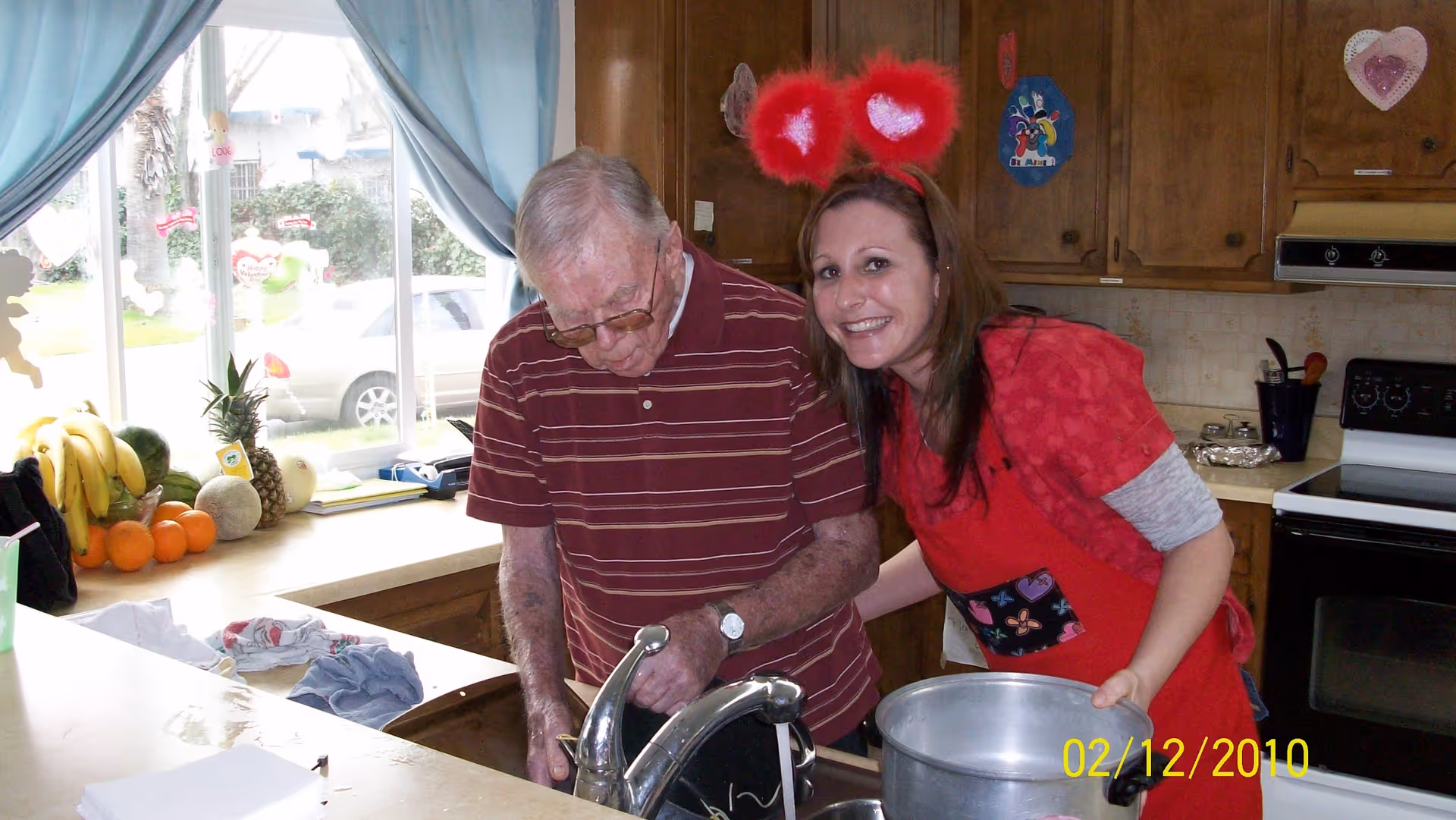 An elderly man and a woman in a kitchen. The man is wearing a striped maroon shirt and glasses, standing at the sink washing dishes. The woman, wearing a red apron and red heart-shaped headband, is smiling and leaning close to the man while holding a large pot. The kitchen has wooden cabinets, a stove, and a window with blue curtains. Various fruits are on the counter near the window.