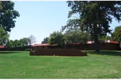 Green lawn with a low brick planter in front of a single-story red-roofed building and tall trees under a blue sky.