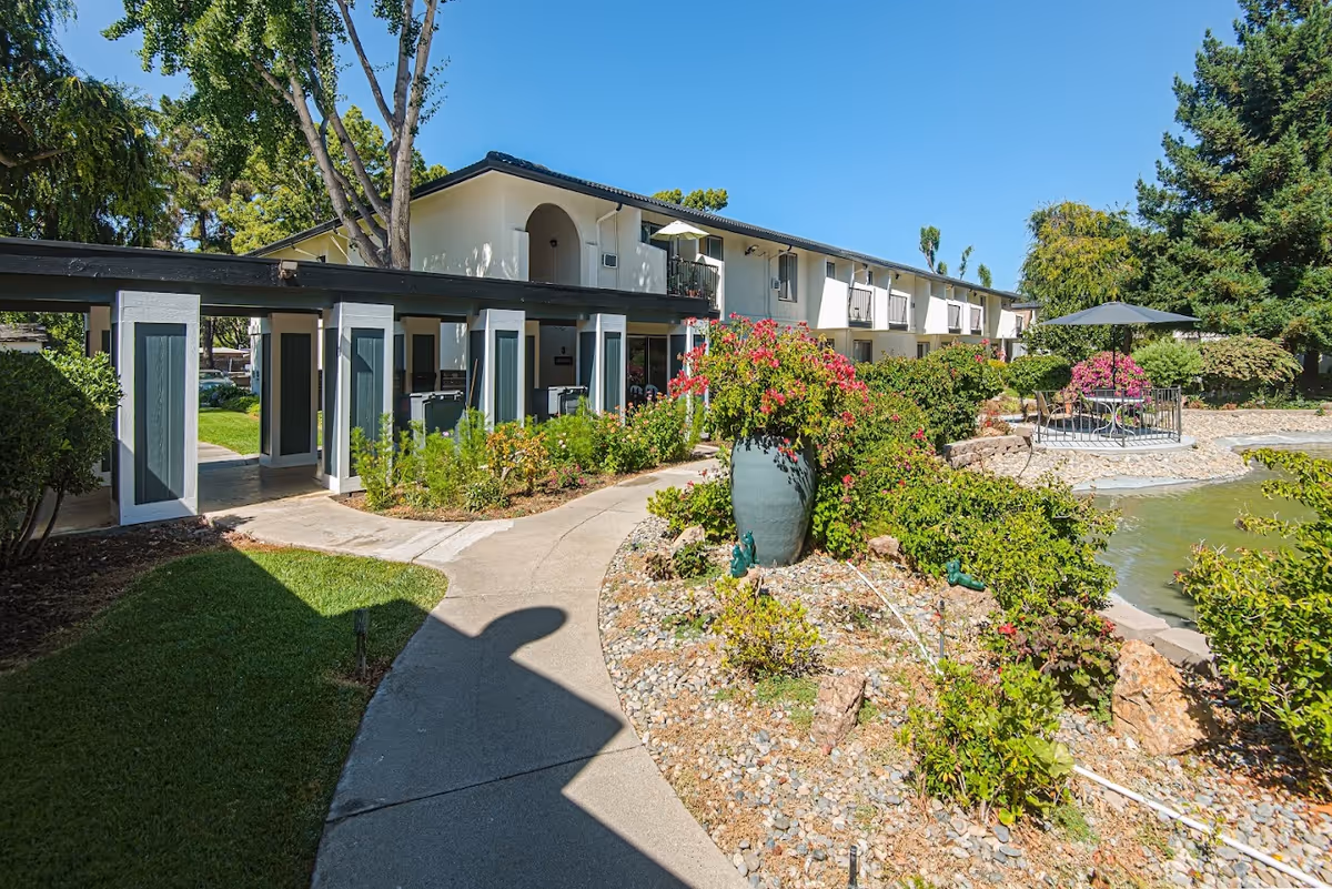 Landscaped courtyard with a winding path, pond and outdoor seating in front of a two-story retirement community building.