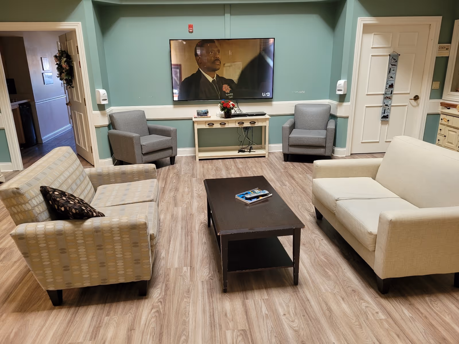A cozy living room area with two armchairs and two sofas arranged around a dark wooden coffee table. A flat-screen TV is mounted on a light green wall above a small white console table with a flower vase and some books. The floor is wood-patterned, and there are two doors visible, one on the left and one on the right.