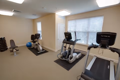 A small fitness room with exercise equipment including a treadmill, elliptical machine, stationary bike, a bench, dumbbells on a rack, and a blue exercise ball. The room has beige walls, carpeted floor, and a large window with blinds letting in natural light.