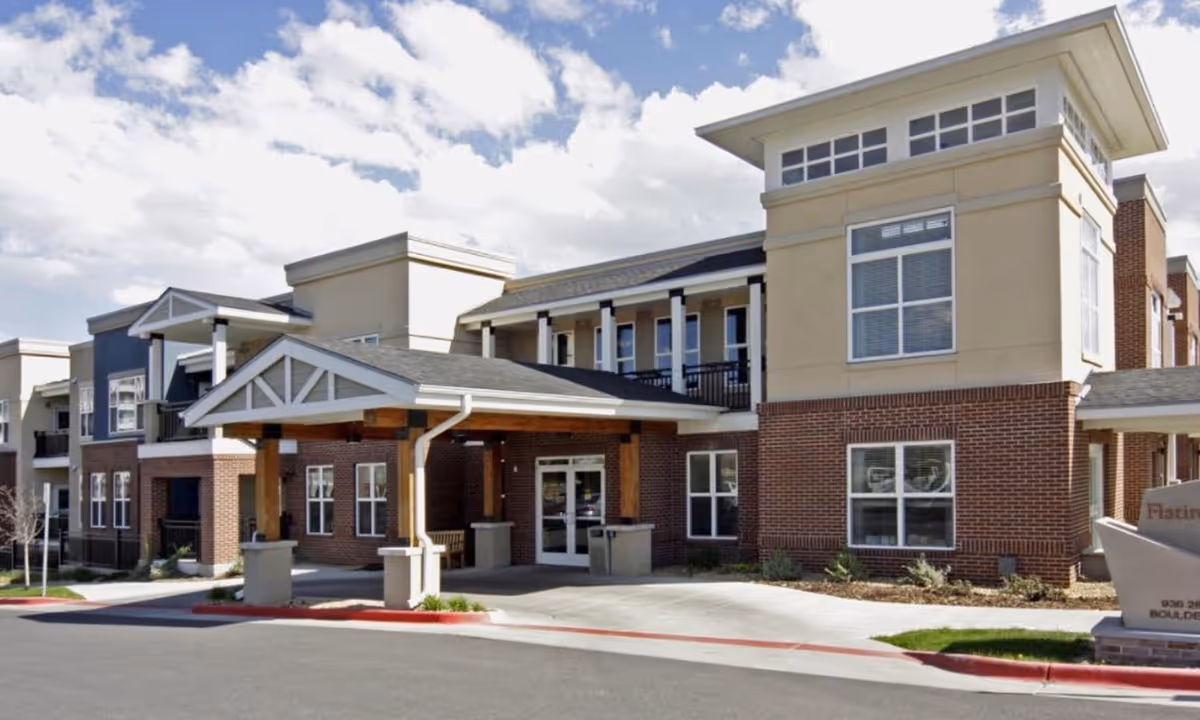 Front entrance of a two-story senior living facility with a covered porte-cochere and brick facade under a partly cloudy sky.