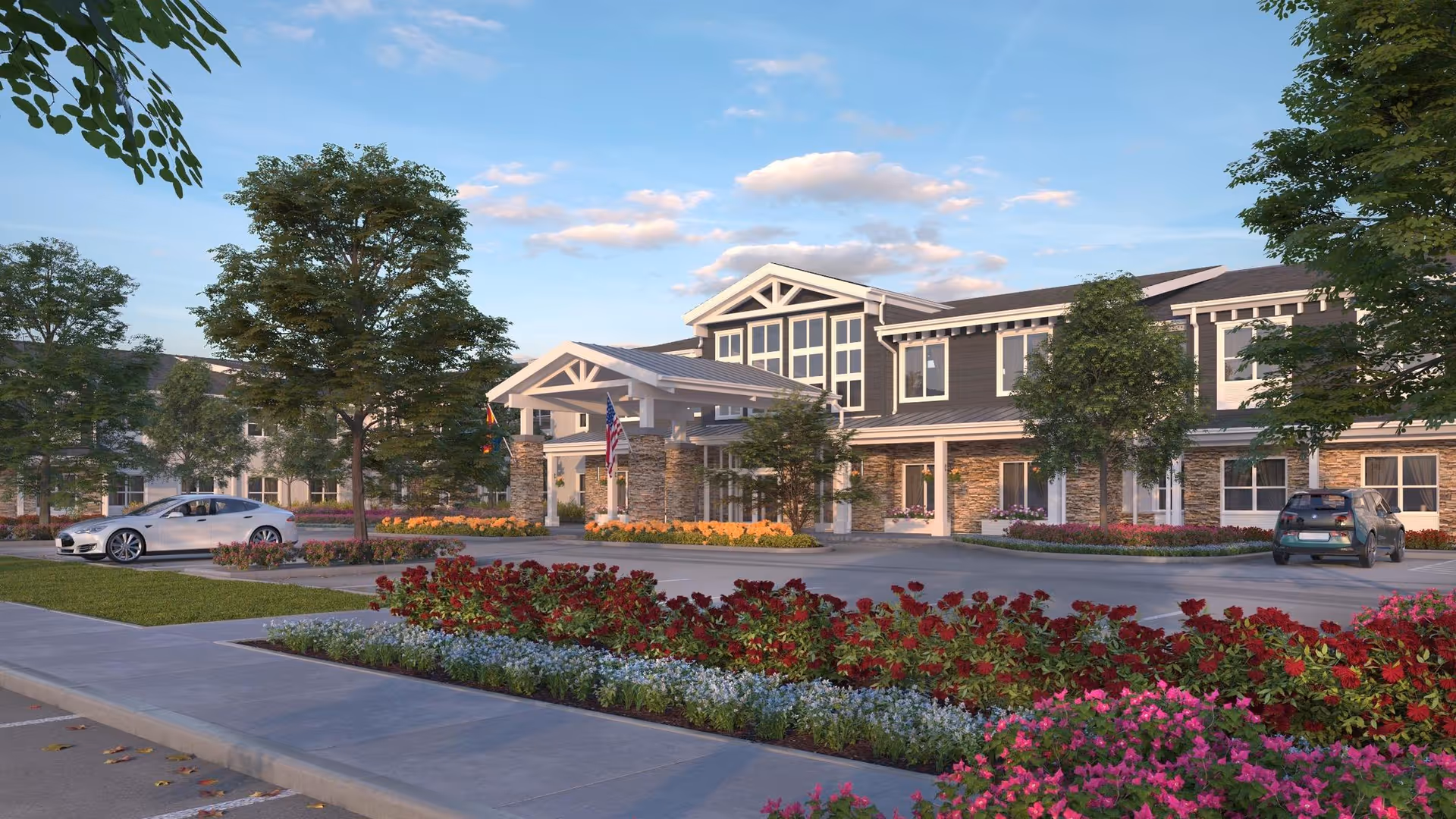 Front exterior of a two-story senior living building with a covered entrance, landscaped flowerbeds, trees, and parked cars under a blue sky.