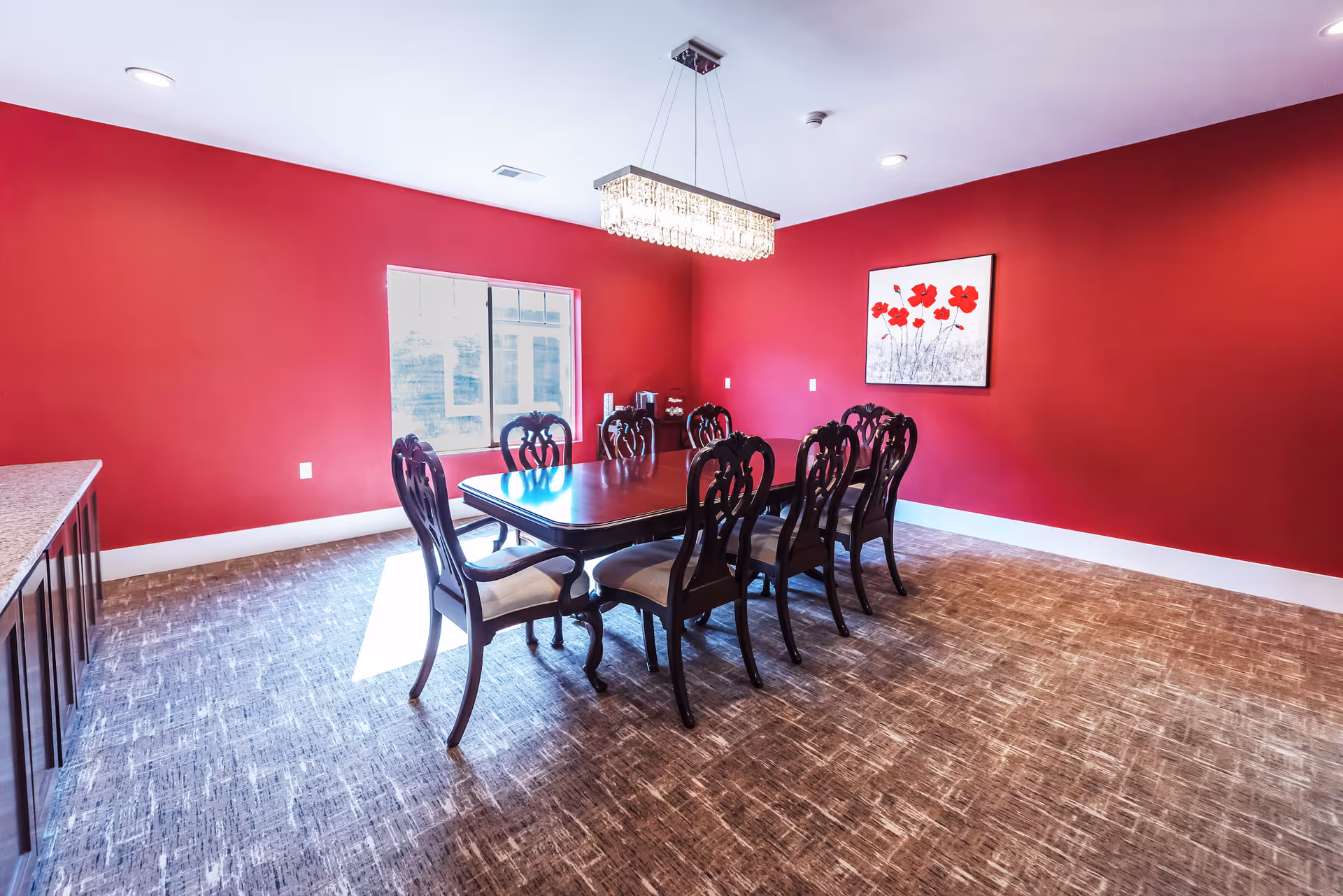 Dining room with a long wooden table and eight chairs, red walls, a window and a chandelier.
