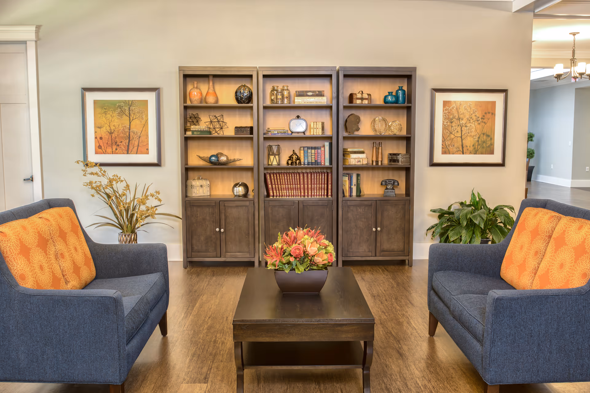 Cozy seating area with two blue armchairs with orange pillows facing a coffee table and a wooden bookshelf filled with books and decorative items.